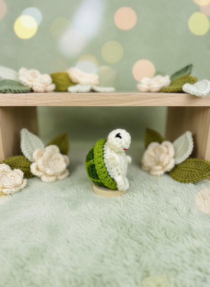 Crochet turtle plushie by Craft-N-Crumble™, surrounded by handmade flowers on a soft green background.