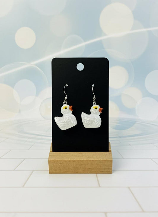 White duck-shaped earrings on a black card with a wooden stand against a white background